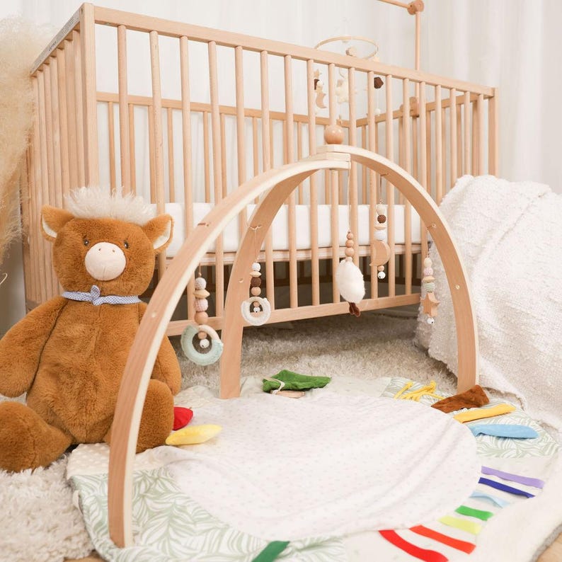 Pode incluir: Um gin&aacute;sio de madeira para beb&eacute;s com brinquedos suspensos, um tapete de brincar macio e um urso de peluche. O gin&aacute;sio tem uma estrutura arqueada com brinquedos de madeira e tecido pendurados. Um ber&ccedil;o de madeira est&aacute; no fundo.