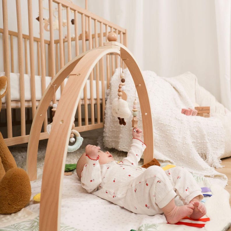 Pode incluir: Um gin&aacute;sio de madeira para beb&eacute;s com brinquedos suspensos. O gin&aacute;sio tem um design de arco curvo e &eacute; feito de madeira de cor clara. Um beb&eacute; com um body branco com estampas de cerejas vermelhas est&aacute; deitado num tapete de brincar.