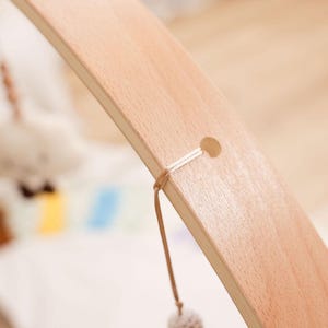 Pode incluir: Close-up de uma estrutura de madeira para m&oacute;bile de beb&ecirc;. A madeira clara tem um acabamento liso e uma forma curva. Um pequeno orif&iacute;cio &eacute; vis&iacute;vel perto do topo, com uma corda presa. Uma pequena bola fofa pende da corda.