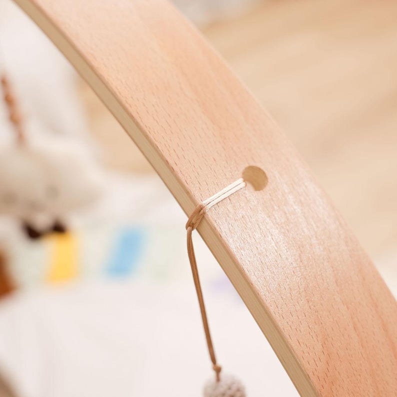 Pode incluir: Close-up de uma estrutura de madeira para m&oacute;bile de beb&ecirc;. A madeira clara tem um acabamento liso e uma forma curva. Um pequeno orif&iacute;cio &eacute; vis&iacute;vel perto do topo, com uma corda presa. Uma pequena bola fofa pende da corda.