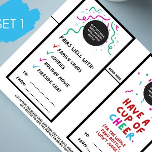 May include: Set of two white wine bottle tags with colorful text and confetti. One tag reads "Pairs well with: Family Chaos, Cookies, Holiday Movie, Fireside Chat." The other says "Have a Cup of Cheer."