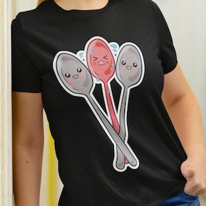 May include: Black t-shirt with a graphic of three cartoon spoons. The spoons have faces and are crossed over each other. The middle spoon is red and has a sad expression. The other two spoons are gray and have happy expressions.