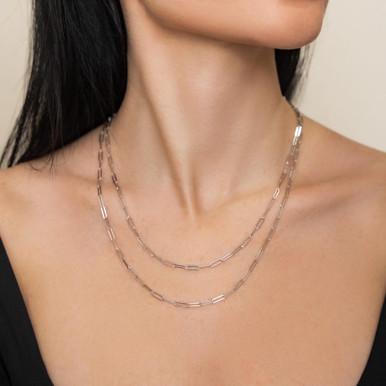 May include: Two silver paperclip chain necklaces. The necklaces are layered, with one resting higher on the neck than the other. The chains are made of small, rectangular links. The background is a neutral colour.