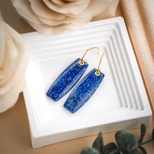 May include: A pair of blue lapis lazuli earrings with gold-colored hoops. The rectangular-shaped stones have a speckled pattern and are displayed on a white, tiered surface. The background includes a cream-colored rose and a beige fabric.