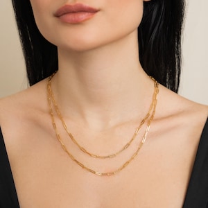 May include: Two gold-coloured paperclip chain necklaces layered on a person's neck. The necklaces are made of rectangular links and are of different lengths. The person is wearing a black top.