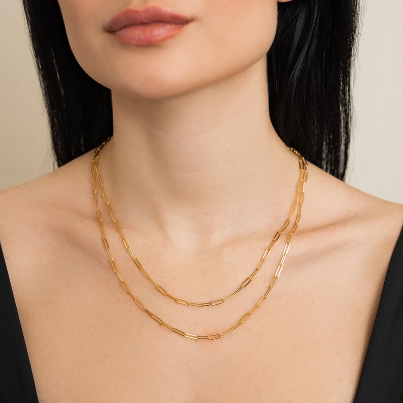 May include: Two gold-coloured paperclip chain necklaces layered on a person's neck. The necklaces are made of rectangular links and are of different lengths. The person is wearing a black top.