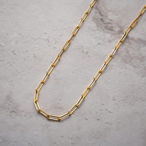 May include: A gold-coloured chain necklace with rectangular links. The necklace is displayed on a light grey surface. The chain is made of a shiny, metallic material and is designed to be worn around the neck.