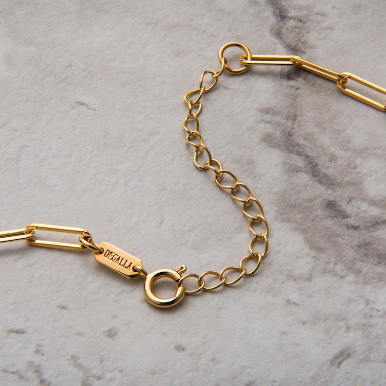 May include: Close-up of a gold necklace with a paperclip chain design. The necklace has a lobster clasp and an extender chain. The brand name "DEGALLA" is engraved on a small rectangular tag.