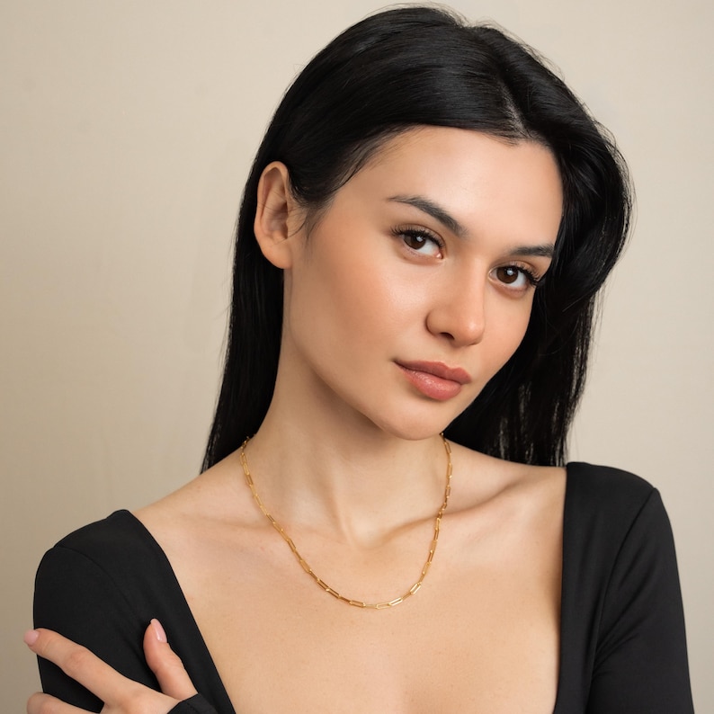 May include: A gold chain necklace featuring rectangular links. The necklace is worn by a person in a black top. The background is a plain, neutral colour.