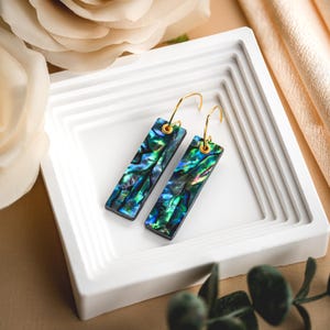 May include: A pair of rectangular earrings with iridescent abalone shells in shades of green, blue, and purple. The earrings have gold-colored hooks and are displayed in a white, tiered tray. The background includes a beige fabric and a white flower.