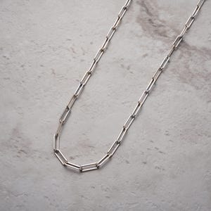 May include: A silver paperclip chain necklace. The necklace has a series of rectangular links, creating a modern and minimalist design. The chain is displayed against a light grey marble surface.