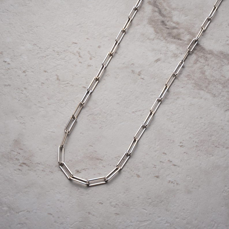 May include: A silver paperclip chain necklace. The necklace has a series of rectangular links, creating a modern and minimalist design. The chain is displayed against a light grey marble surface.
