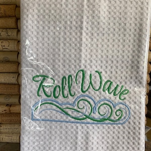 May include: White waffle weave kitchen towel with green and blue embroidered waves and the text "Roll Wave".