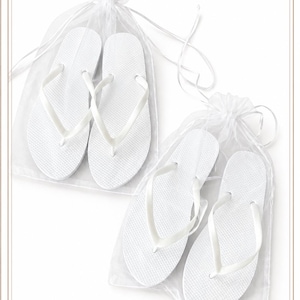 May include: Two pairs of solid white flip-flops, each pair individually packaged in a sheer white organza drawstring bag. The flip-flops feature a textured footbed and smooth, thin straps, presented in a clean, minimalist arrangement.