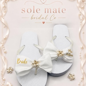 May include: A pair of white flip-flops featuring large satin bows adorned with pearl and rhinestone flower centres. The left sandal has the word Bride in gold script, and the right sandal has Mrs. Holmes in gold script. The text sole mate bridal Co is above.