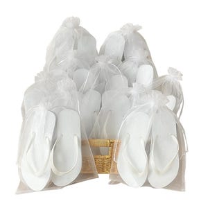 May include: A collection of white flip-flops, each individually packaged in a sheer white organza drawstring bag. The flip-flops are arranged together in a light-coloured woven basket, creating a uniform display of multiple pairs of footwear.