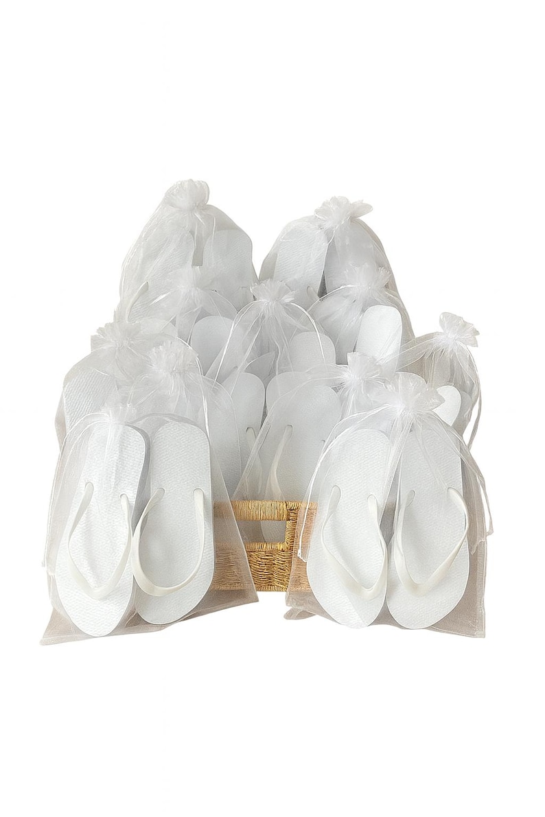May include: A collection of white flip-flops, each individually packaged in a sheer white organza drawstring bag. The flip-flops are arranged together in a light-coloured woven basket, creating a uniform display of multiple pairs of footwear.