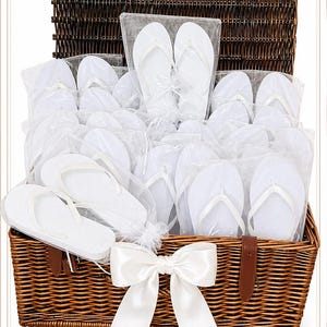 May include: A large brown woven wicker basket filled with multiple pairs of white flip-flops, each individually packaged in clear plastic bags. The basket features a hinged lid and is decorated with a large, white satin ribbon bow on the front.