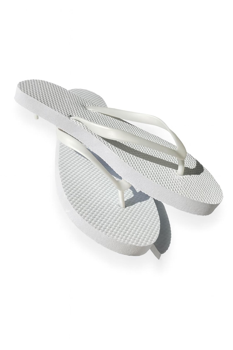 May include: A pair of solid white flip-flops featuring a textured footbed with a raised dot pattern and smooth, Y-shaped straps. The sandals are constructed from a lightweight foam material, showcasing a simple design with no additional patterns or colours.
