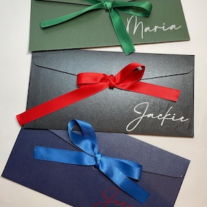 May include: Three coloured envelopes with satin ribbons. The top envelope is dark green with a green ribbon and the name "Maria" in white script. The middle envelope is black with a red ribbon and the name "Jackie" in white script. The bottom envelope is navy blue with a blue ribbon and the name "Jessica" in red script.