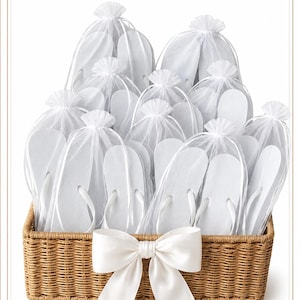 May include: A rectangular woven wicker basket containing multiple pairs of white flip-flops, each individually packaged in a sheer white organza drawstring bag. The basket is decorated with a large, centred white satin ribbon bow on the front.