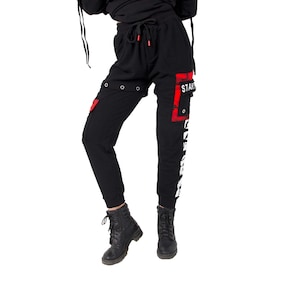 Może przedstawiać: Czarne spodnie dresowe z czerwono-białym nadrukiem graficznym. Spodnie dresowe mają sznurek w pasie i boczne kieszenie. Nadruk zawiera słowo "START" i inne elementy graficzne.