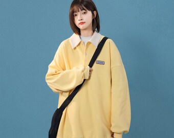 May include: A yellow long-sleeved collared sweatshirt with a white collar and a small patch on the left chest. The sweatshirt has a relaxed fit and is being worn by a person with short brown hair.