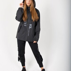 May include: Black hooded jacket with the text "KIL" and "AVEN" printed on the front. The jacket has a zipper closure and two pockets. The model is wearing black cargo pants and black sneakers.
