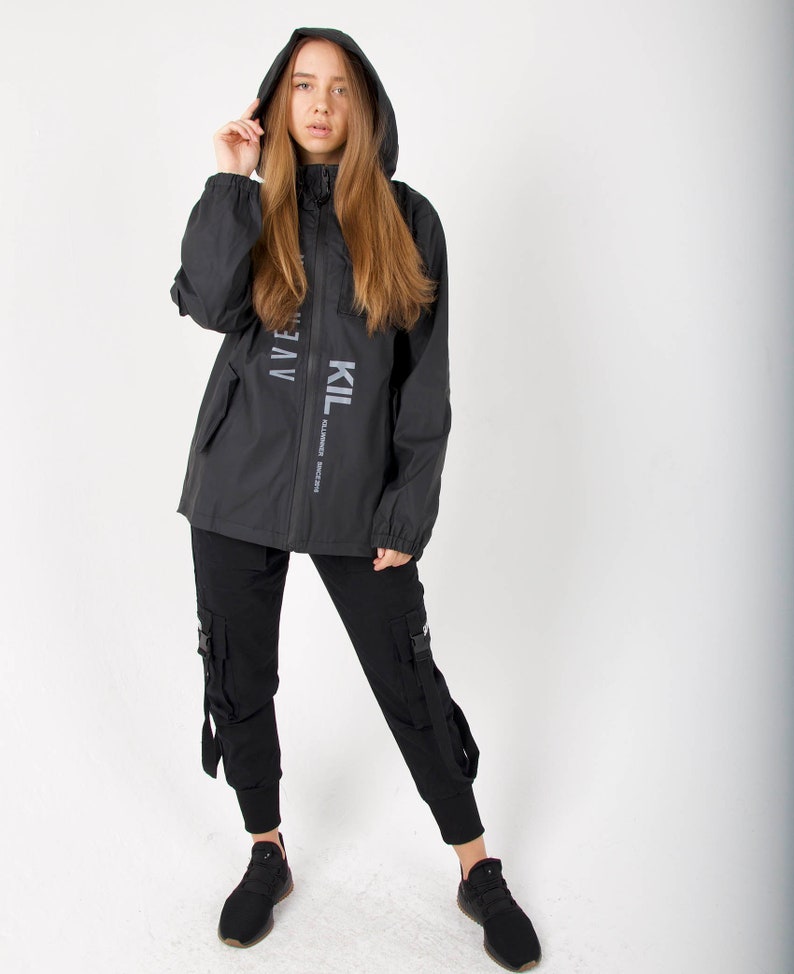 May include: Black hooded jacket with the text "KIL" and "AVEN" printed on the front. The jacket has a zipper closure and two pockets. The model is wearing black cargo pants and black sneakers.