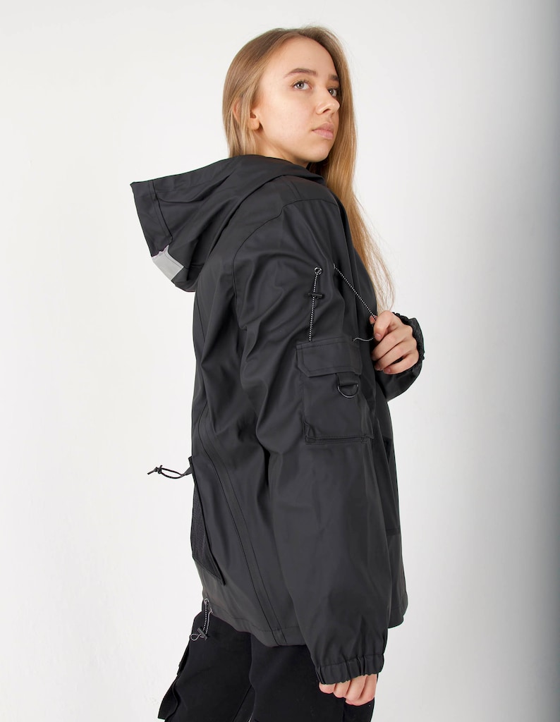 May include: A black hooded jacket with a drawstring waist and a reflective detail on the hood. The jacket has a zipper closure and a large pocket on the left sleeve.