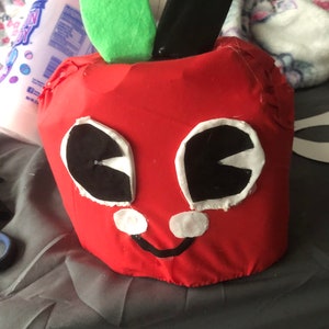 May include: A red apple-shaped plush toy with black eyes, white accents, and a black smile. The apple has a green felt leaf and a black felt stem.