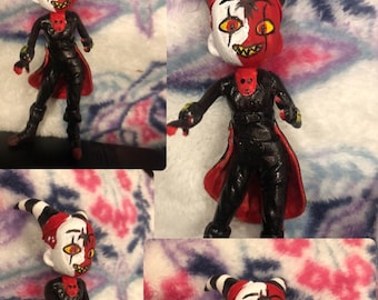 May include: A handmade figurine of a character with a red and white face, black and red clothing, and black horns. The figurine is painted with details, including a spider on the face.