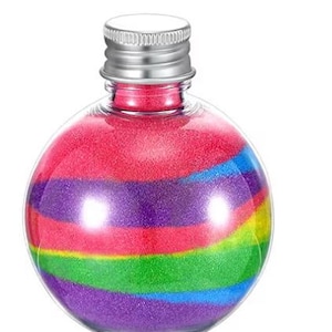 May include: A clear, round glass bottle filled with layers of colorful sand. The sand is arranged in horizontal bands of red, purple, green, yellow, and blue. The bottle has a silver metal cap.