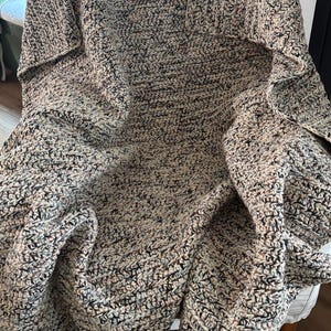 May include: A chunky crochet blanket featuring a variegated yarn pattern with blended tones of cream, tan, brown, and black. The textile has a thick, textured stitch pattern and is draped over a chair, showcasing its soft, heavy, and woven construction.