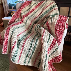 May include: A handmade crochet blanket draped over a chair. The blanket features a cream-colored base with diagonal stripes of red and green. The texture of the yarn is visible, creating a cozy and inviting aesthetic. The blanket is a decorative home textile.