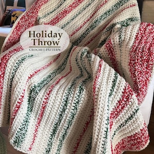 May include: A crochet throw blanket featuring a textured pattern of vertical stripes in cream, red, and dark green. The blanket is draped over a chair and includes an oval overlay with the text Holiday Throw CROCHET PATTERN printed in dark brown.