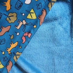 Könnte beinhalten: Ein blauer Stoff mit einem Muster aus Füchsen, Rehen, Pilzen und Bäumen. Der Stoff ist in der Mitte gefaltet, wobei die blaue Seite unten und die gemusterte Seite oben zu sehen ist.