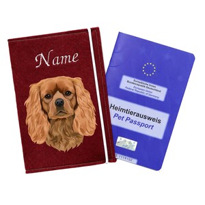 Könnte beinhalten: Ein roter Filz-Reisepasshalter mit einem weißen bestickten Namensschild und einer bunten Illustration eines Cavalier King Charles Spaniels. Der Halter liegt neben einem blauen EU-Heimtierausweis mit dem Text "Heimtierausweis Pet Passport" und einem EU-Sternensymbol.