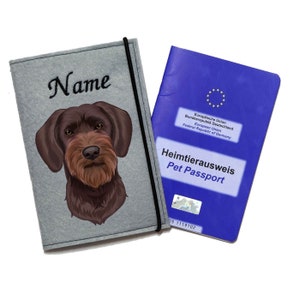 Könnte beinhalten: Grauer Filz-Reisepasshalter mit einem gestickten braunen Hunde-Kopf auf der Vorderseite. Der Text "Name" ist über dem Hunde-Kopf gestickt. Der Halter befindet sich neben einem blauen Heimtierausweis mit dem Text "Heimtierausweis Pet Passport" und "Europäische Union Bundesrepublik Deutschland Federal Republic of Germany European Union".