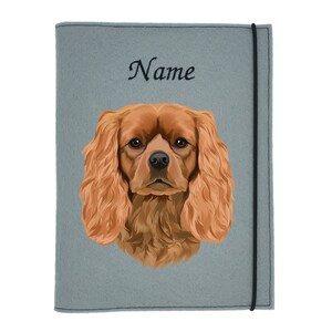 Könnte beinhalten: Grauer Filzordner mit schwarzem Gummibandverschluss. Der Ordner hat ein gesticktes Bild eines Cavalier King Charles Spaniel Hundes mit dem Wort "Name" gestickt über dem Hund.