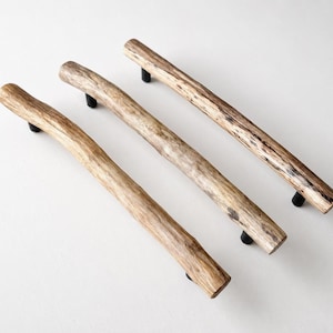 Könnte beinhalten: Drei rustikale Holzschrankgriffe mit schwarzer Hardware. Die Griffe bestehen aus natürlich aussehenden Ästen, die verschiedene Braun- und Beigetöne aufweisen. Die Hardware ist ein einfaches, rundes, schwarzes Design.