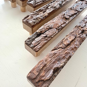 Op de afbeelding: Close-up van verschillende houten meubelgrepen met een natuurlijke boomschors textuur op het bovenoppervlak. De grepen zijn rechthoekig met een lichtgekleurde houten basis en een donkerbruine, getextureerde schorsafwerking. De grepen zijn gerangschikt op een wit oppervlak.