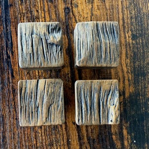 Puede incluir: Cuatro tiradores cuadrados de madera con un aspecto envejecido y texturizado. Los tiradores son una mezcla de marrón y gris, con vetas y ranuras de madera visibles. Están dispuestos en forma cuadrada, sugiriendo un juego para muebles.