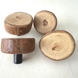 May include: Four wooden cabinet knobs with a natural, rustic aesthetic. The knobs are made from cross-sections of tree branches, showcasing the wood's grain and bark. One knob is attached to a black metal stem.