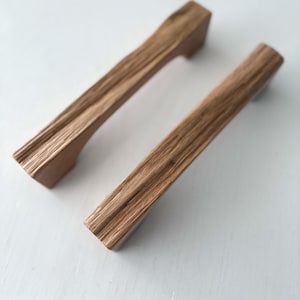 Puede incluir: Dos tiradores de armario de madera con un acabado de vetas naturales. Los tiradores son rectangulares con un asa ligeramente curvada y miden aproximadamente 15 cm de largo. Están diseñados para un agarre fácil y añaden un toque rústico a cualquier mueble.