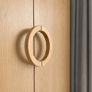 May include: A light-colored wooden cabinet door featuring a circular, two-piece wooden handle mounted over the vertical seam. The wood grain is visible on both the flat door surface and the curved, ring-shaped handle, showing a natural finish.