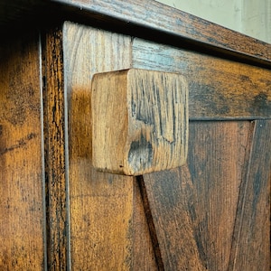 Könnte beinhalten: Nahaufnahme einer Holztür mit einem quadratischen, strukturierten Holzgriff. Der Schrank hat eine rustikale, abgenutzte Oberfläche mit dunkelbraunen und schwarzen Tönen und sichtbarer Holzmaserung.
