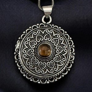 Pode incluir: Um colar com pingente prateado com um design circular intrincado. O pingente apresenta uma pedra central redonda de cor âmbar e padrões de filigrana detalhados. O colar pende de uma fina corrente prateada sobre um fundo azul escuro.