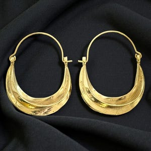 Peut inclure: Une paire de boucles d'oreilles créoles dorées. Chaque boucle d'oreille présente un design en forme de croissant avec un intérieur creux. Les boucles d'oreilles sont présentées sur un fond en tissu noir, mettant en valeur leur éclat métallique.