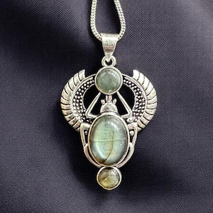 Peut inclure: Un collier pendentif argenté avec un motif de scarabée égyptien. Le pendentif comporte trois pierres précieuses ovales vert pâle et des détails d'ailes. Le collier est présenté sur un tissu gris foncé.